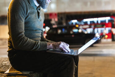 Freelancer using laptop sitting on bench in city at night - WPEF07922