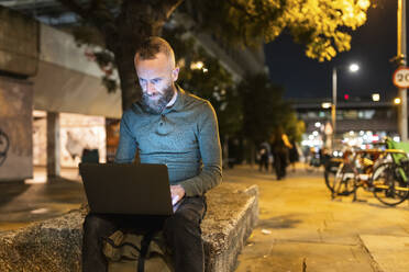 Freelancer working on laptop in city at night - WPEF07923