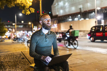 Nachdenklicher Freiberufler sitzt mit Laptop in der nächtlichen Stadt - WPEF07924