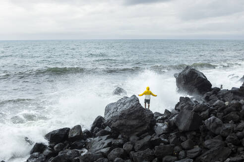 Carefree young man standing on rocks near splashing waves in front of sea - WVF02045