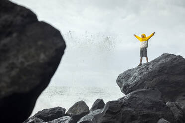 Man with arms raised standing on rock near splashing waves of sea - WVF02046