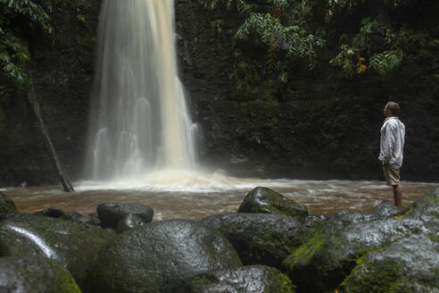 Junger Mann steht auf Felsen in der Nähe eines Wasserfalls im Regenwald - WVF02071