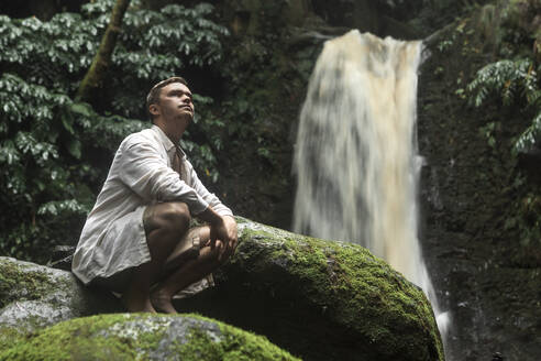 Nachdenklicher junger Mann, der auf einem Felsen in der Nähe eines Wasserfalls kauert - WVF02076