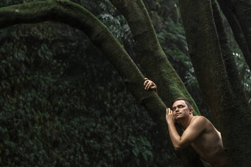 Shirtless man with eyes closed leaning on tree trunk - WVF02078