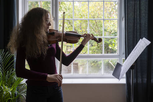 Mädchen spielt Geige vor einem Fenster zu Hause - NJAF00685