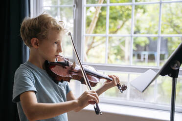 Junge übt Geige mit Notenblättern in der Nähe des Fensters zu Hause - NJAF00689