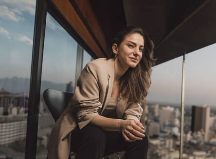 Smiling businesswoman sitting on chair near window in office - MFF09459