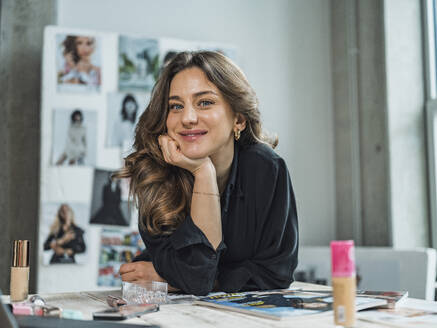 Smiling freelancer sitting with beauty products at desk in studio - MFF09481