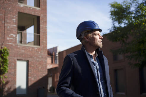 Thoughtful architect wearing hardhat in front of buildings - KNSF10009