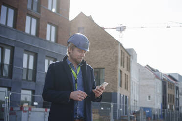 Architekt trägt Schutzhelm und benutzt Mobiltelefon in der Nähe von Gebäuden - KNSF10011