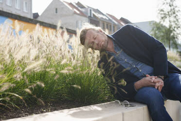 Businessman with eyes closed leaning on grass in front of houses - KNSF10025