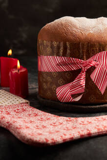 A beautifully presented panettone bread wrapped with a striped red ribbon, accompanied by a decorative fabric with festive patterns, set amidst ambient candlelight. - ADSF49836