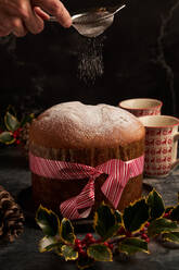 Close-up of a hand sprinkling powdered sugar over a festive panettone, surrounded by holiday decorations and ceramic mugs - ADSF49838
