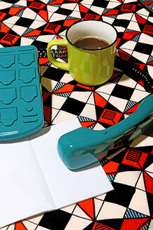 High angle of turquoise vintage telephone alongside a green mug filled with coffee, placed on a striking geometric-patterned backdrop near blank white paper - ADSF49840