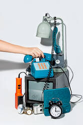 Ein altes Telefon in der Hand vor einem weißen Hintergrund, in dem ein alter Fernseher, ein Wecker, eine Schreibtischlampe und einige andere Retro-Geräte zu sehen sind - ADSF49843