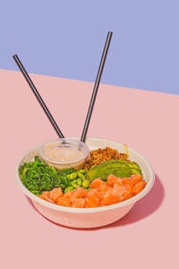 High angle of poke bowl filled with fresh salmon, green edamame beans, crunchy seaweed salad, creamy avocado, and crunchy granola, accompanied by a light dipping sauce and chopsticks, against a dual-toned pastel background - ADSF49844