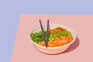 High angle of poke bowl filled with fresh salmon, green edamame beans, crunchy seaweed salad, creamy avocado, and crunchy granola, accompanied by a light dipping sauce and chopsticks, against a dual-toned pastel background - ADSF49845