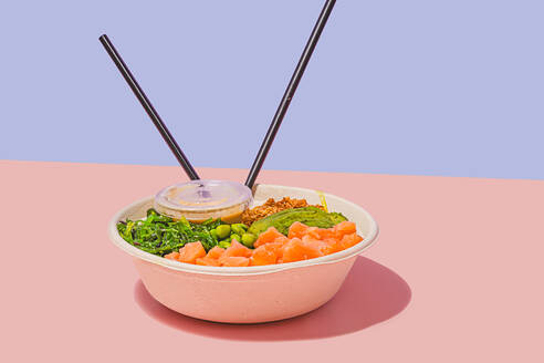 Poke bowl filled with fresh salmon, green edamame beans, crunchy seaweed salad, creamy avocado, and crunchy granola, accompanied by a light dipping sauce and chopsticks, against a dual-toned pastel background - ADSF49846