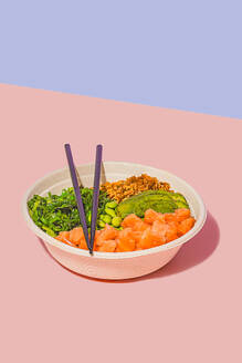 High angle of poke bowl filled with fresh salmon, green edamame beans, crunchy seaweed salad, creamy avocado, and crunchy granola, accompanied by a light dipping sauce and chopsticks, against a dual-toned pastel background - ADSF49847