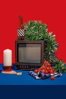 Vintage television set surrounded by an array of objects, including a bottle with a striped label, fresh grapes on a checkered cloth, a white candle and green tinsel, all set against a red backdrop on a blue table - ADSF49848