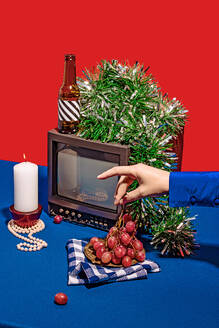 High angle of vintage television set surrounded by an array of different objects on blue table against red background, while a crop anonymous person picking a grape from the checkered cloth-covered stack - ADSF49849