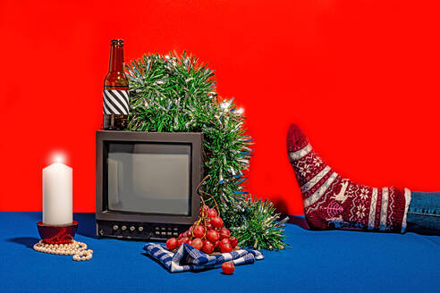 Vintage television set surrounded by an array of objects, including a bottle with a striped label, fresh grapes on a checkered cloth, a white candle and green tinsel, all set against a red backdrop on a blue table with a crop foot with festively decorated sock - ADSF49850