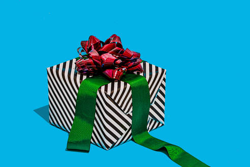 Striped black and white gift box adorned with a shiny red bow and a green ribbon placed against a vibrant turquoise background - ADSF49851
