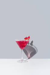 Glass filled with red pomegranate cocktail served with pomegranate seeds against a muted gray backdrop, casting a soft shadow - ADSF49890