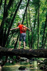 Adventurous man in a bright red t-shirt carefully hiking on a fallen log on green forest stream, looking ahead. - ADSF49898