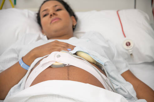 Close-up of an expectant mother in a hospital bed, touching her belly with a fetal heart rate monitor strapped around her - ADSF49905