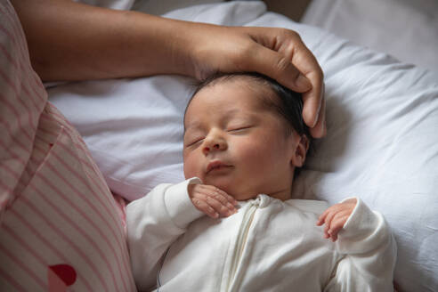 A gentle hand caresses the head of a sleeping newborn baby, dressed in a white onesie on soft bedding. - ADSF49906