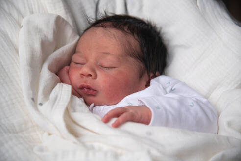 A newborn baby rests peacefully on white bedding, clad in a soft white onesie with hands delicately posed - ADSF49907