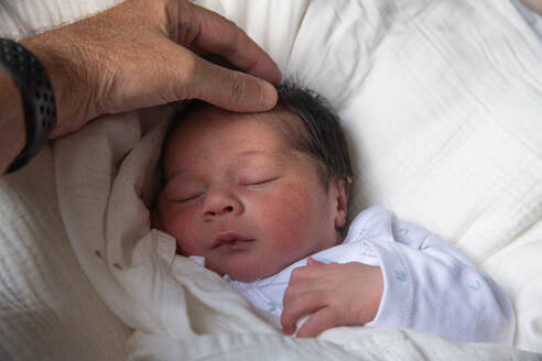 A gentle hand caresses the head of a sleeping newborn baby, dressed in a white onesie on soft bedding. - ADSF49908