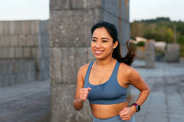 A smiling Latin American woman running outdoors in workout attire, embodying an active lifestyle and positive energy. - ADSF49916