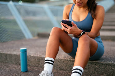 A young Latin American woman sits on steps during a workout break, checking her smartphone with a water bottle nearby. - ADSF49920