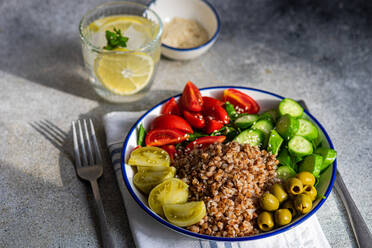 High angle of healthy lunch bowl with boiled organic buckwheat, fresh cucumber and tomato, and fermented tomato and olives served with glass of pure water with lemon, ice and mint against blurred gray surface - ADSF49923