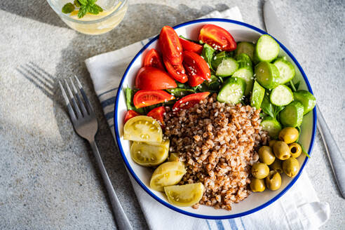 Top view of healthy lunch bowl with boiled organic buckwheat, fresh cucumber and tomato, and fermented tomato and olives served with glass of pure water with lemon, ice and mint against blurred gray surface - ADSF49925