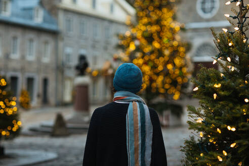 A person in a winter hat stands facing a twinkling Christmas tree in an urban square, creating a cozy holiday atmosphere in Quebec, Canada - ADSF49944