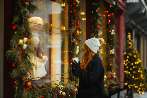 Eine junge Frau mit weißer Strickmütze und schwarzem Mantel hält eine Kaffeetasse in der Hand, während sie eine festliche, mit Lichtern und Dekorationen geschmückte Weihnachtsauslage in Quebec, Kanada, betrachtet - ADSF49945