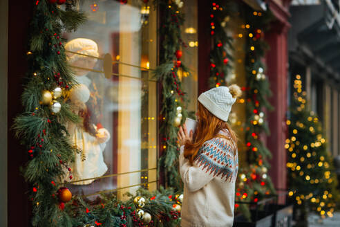 A woman in a warm sweater and beanie enjoys a festive Christmas window display while holding a cup in Quebec, Canada - ADSF49948