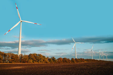 A tranquil scene showcasing a row of wind turbines against a vivid blue sky, with autumnal foliage and a freshly plowed field in the foreground. - ADSF49949