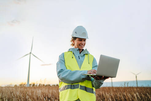 Smiling female engineer in safety vest and hardhat using a laptop while standing at a wind farm field at sunset. - ADSF49956