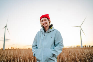 A cheerful young woman wearing a red beanie and a light blue jacket, with a backdrop of wind turbines in a field during the golden hour. - ADSF49959