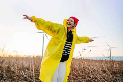 A joyful individual in a yellow raincoat standing in a field during sunset, expressing freedom and happiness by wind turbines. - ADSF49961