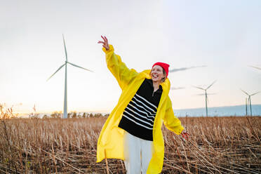 Joyful young woman in a vibrant yellow raincoat and red beanie is dancing in a field during sunset with wind turbines in the background. - ADSF49963