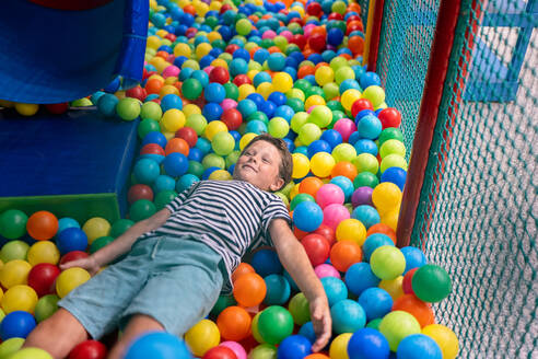 A joyful young boy lies back in a sea of vibrant multicolored plastic balls at a children's indoor play area, depicting fun and playfulness. - ADSF49972