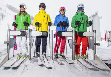 Four skiers in vibrant jackets wait on a ski lift, ready for a day on the slopes, with snow-covered mountains in the background. - ADSF49975