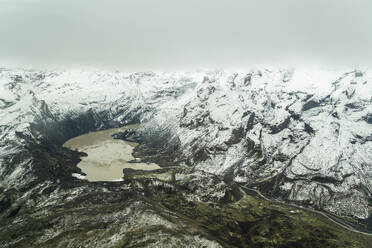 Aerial view of a rugged mountains blanketed in snow with a semi-frozen lake nestled within, under a cloudy sky - ADSF49979