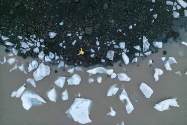 From below featuring person in yellow jacket lying down standing on volcanic black sand beach surrounded by scattered white ice chunks in Vatnajokull National Park, Iceland - ADSF49986