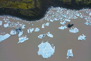 Aerial shot of numerous white ice pieces floating on a dark brown glacial river by the shoreline contrasting with rugged terrain in Vatnajokull National Park - ADSF49988